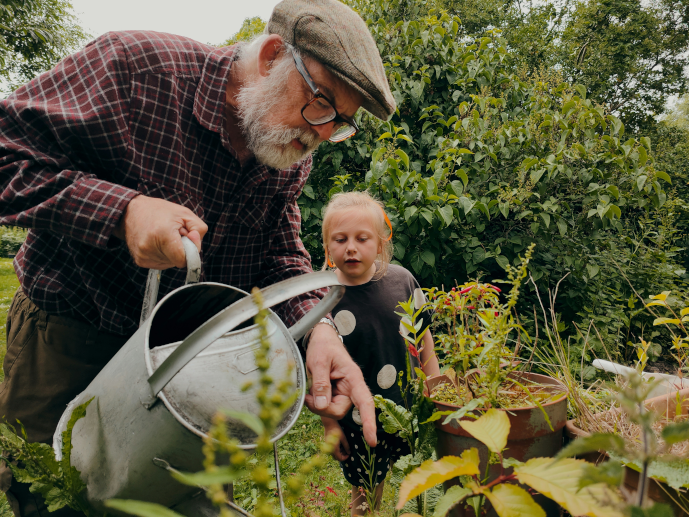 a man holding a watering can points at a plant in front of a young girl